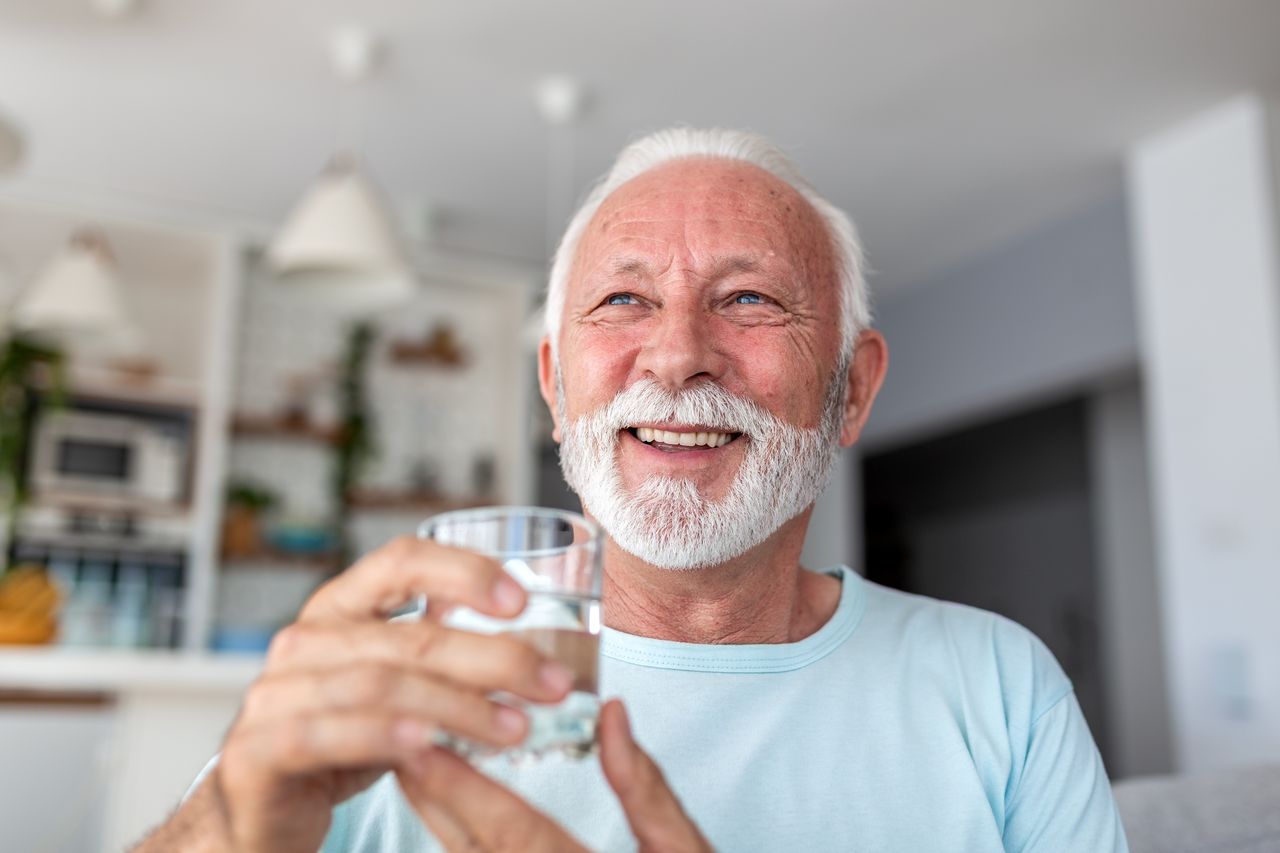 Smiling older man drinking a glass of water at home, representing the public health debate over removing fluoride from community water supplies